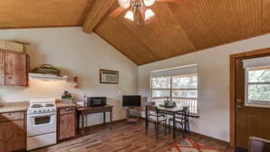 A compact kitchen with a stove, microwave, and wooden cabinets. There is a dining area with a table and chairs, and a window with a view of trees.