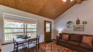 A cozy living room with a wooden ceiling, a brown leather sofa, a dining table with chairs, and a large window with a view of trees.
