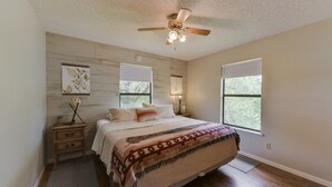 Ein Schlafzimmer mit Bett, Deckenventilator, Nachttisch, Fenster und einem an der Wand hängenden Kunstwerk.