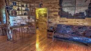 A cabin interior with a wooden floor, a sofa, a dining table with chairs, and shelves with various items.