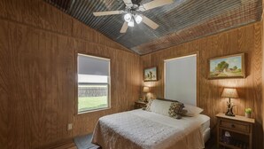 Un dormitorio con paneles de madera, una cama con colcha color crema, dos mesitas de noche con lámparas y un ventilador de techo.