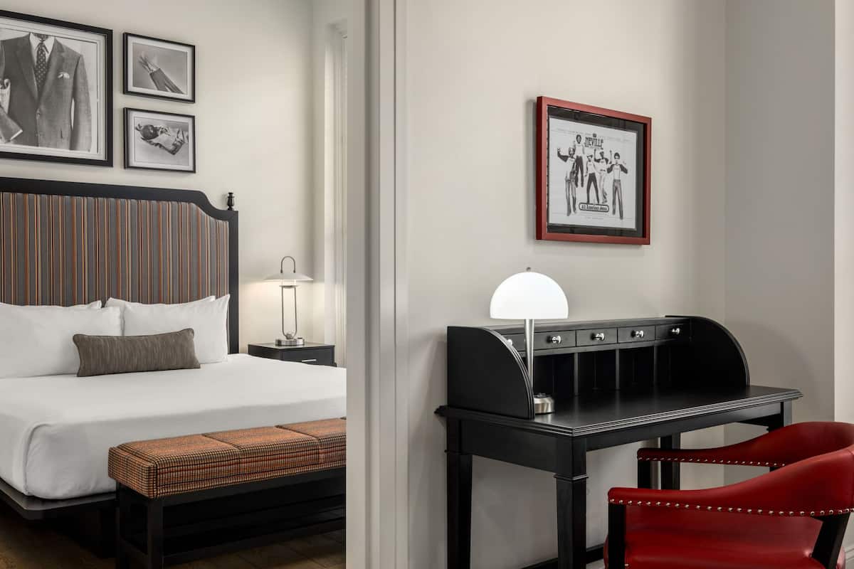 Ein Hotelzimmer mit Bett, Nachttisch, Lampe, Schreibtisch, Stuhl und gerahmten Bildern an der Wand.