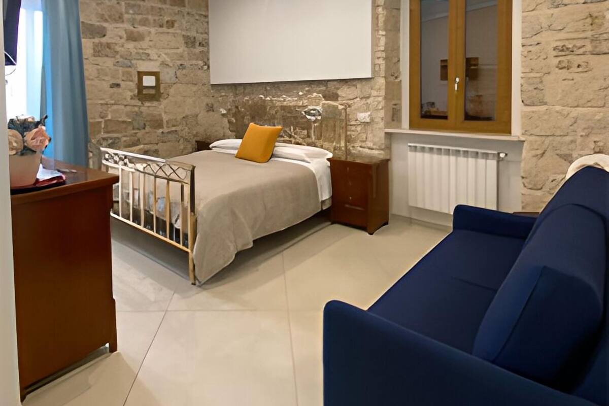 Une chambre moderne avec un mur en pierre, un lit agrémenté d’un coussin beige et orange, un canapé bleu et une table de chevet en bois.