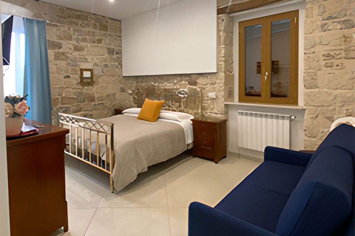 A modern bedroom with a stone wall, a bed with a beige and orange pillow, a blue sofa, and a wooden nightstand.