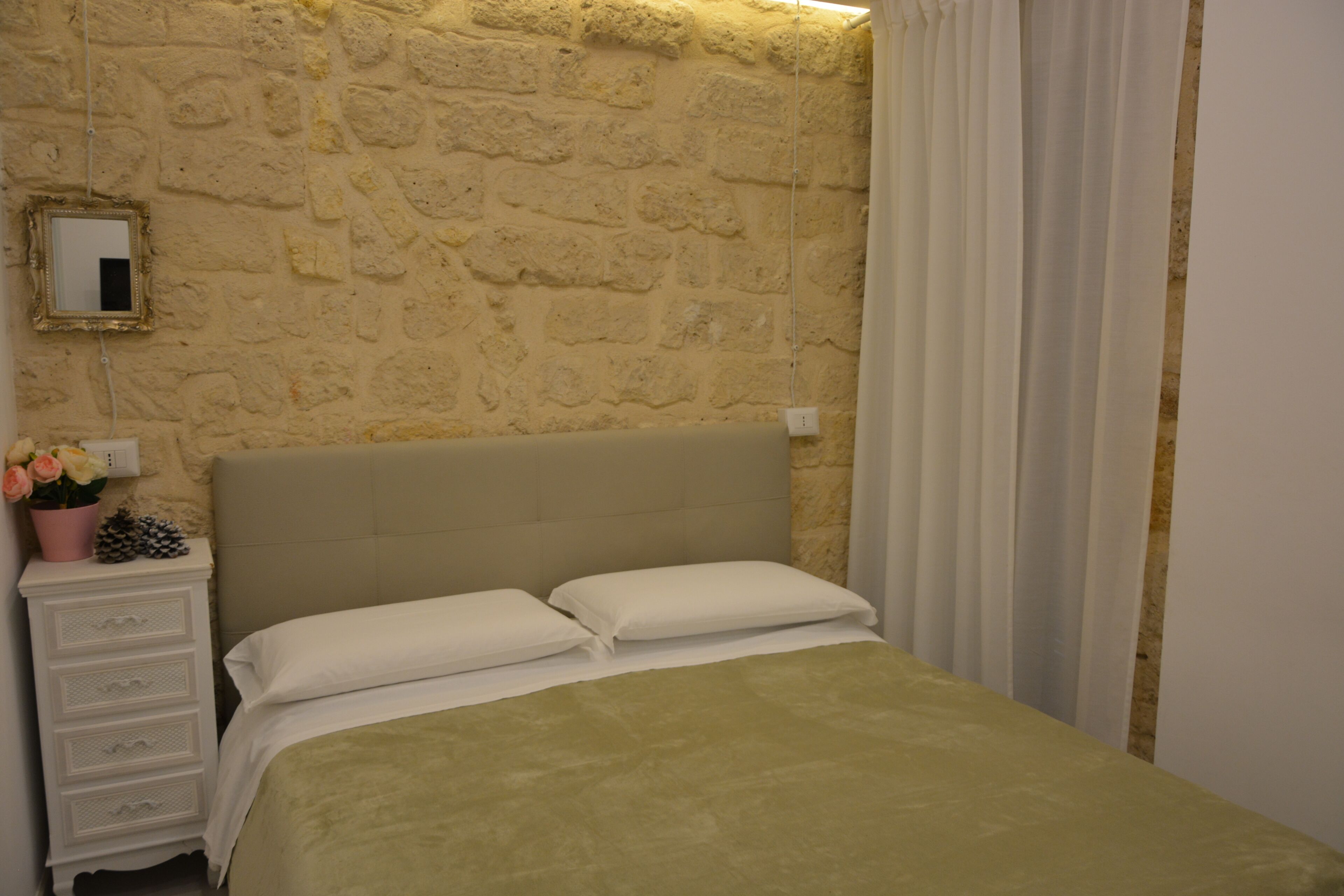 Un dormitorio con pared de piedra, cama con almohadas blancas, mesita de noche con espejo y cómoda.