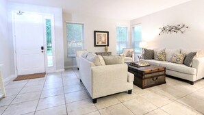 A living room with a beige sofa, a coffee table, and a chessboard. There is a door and a window with blinds.