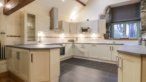 A modern kitchen with white cabinetry, a central island, a built-in oven, and a stone accent wall. - The Barn by Lyons (Talybont)