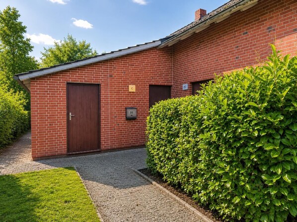 A red brick building with a brown door and two windows, a hedge, and a gravel path.