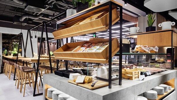 Un café moderno con un mostrador central, sillas de madera y una variedad de alimentos a la vista.