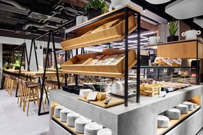 A modern cafe with a central counter, wooden chairs, and a variety of food items displayed. - H2 Hotel Saarbrücken (Saarbrücken)