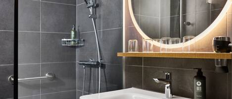 Un baño moderno con un lavabo blanco, un espejo redondo con luz integrada, un cabezal de ducha manual y una botella de jabón para manos.