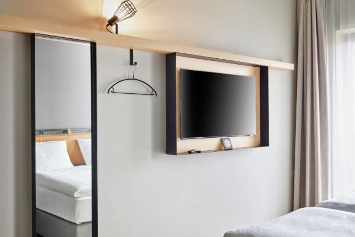 A hotel room with a bed, a large mirror, a television mounted on the wall, and a hanging light fixture.