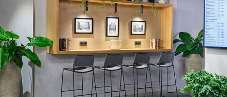 Zona de bar con estante de madera que muestra fotografías enmarcadas, taburetes negros y plantas en macetas.