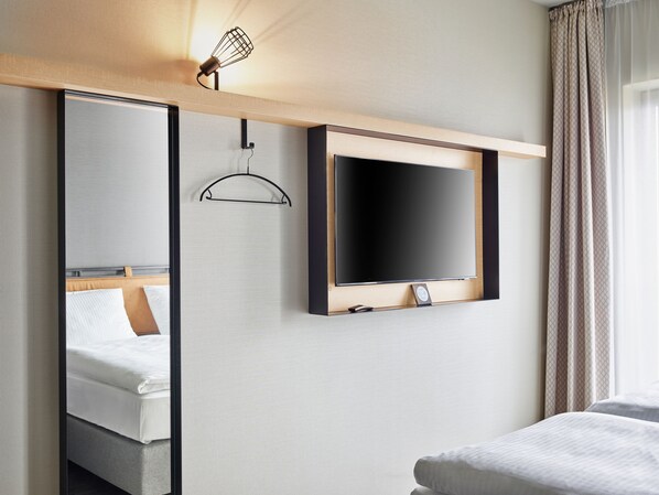 A hotel room with a large mirror, a flat-screen TV mounted on the wall, a hanging coat rack, and a bed with white bedding.