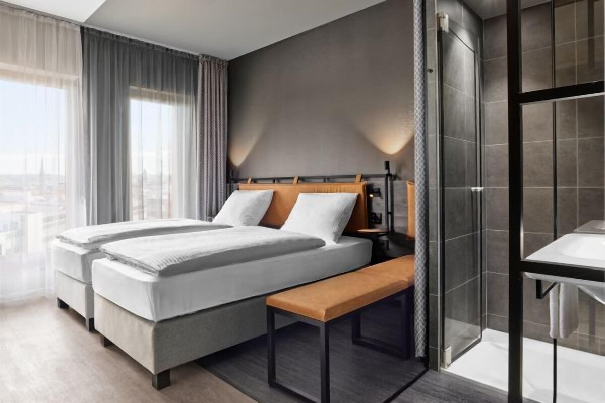 A modern hotel room with a large bed, a bench, a bedside table, and a shower area.