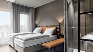 Una habitación de hotel moderna con una cama grande, un banco, una mesita de noche, un espacio ducha y vistas a la ciudad.