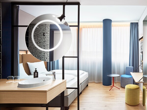 A modern hotel room with a bed, a round mirror, a bedside table with a sink, and a small round table. - Hotel Wien-Schönbrunn (Vienna)