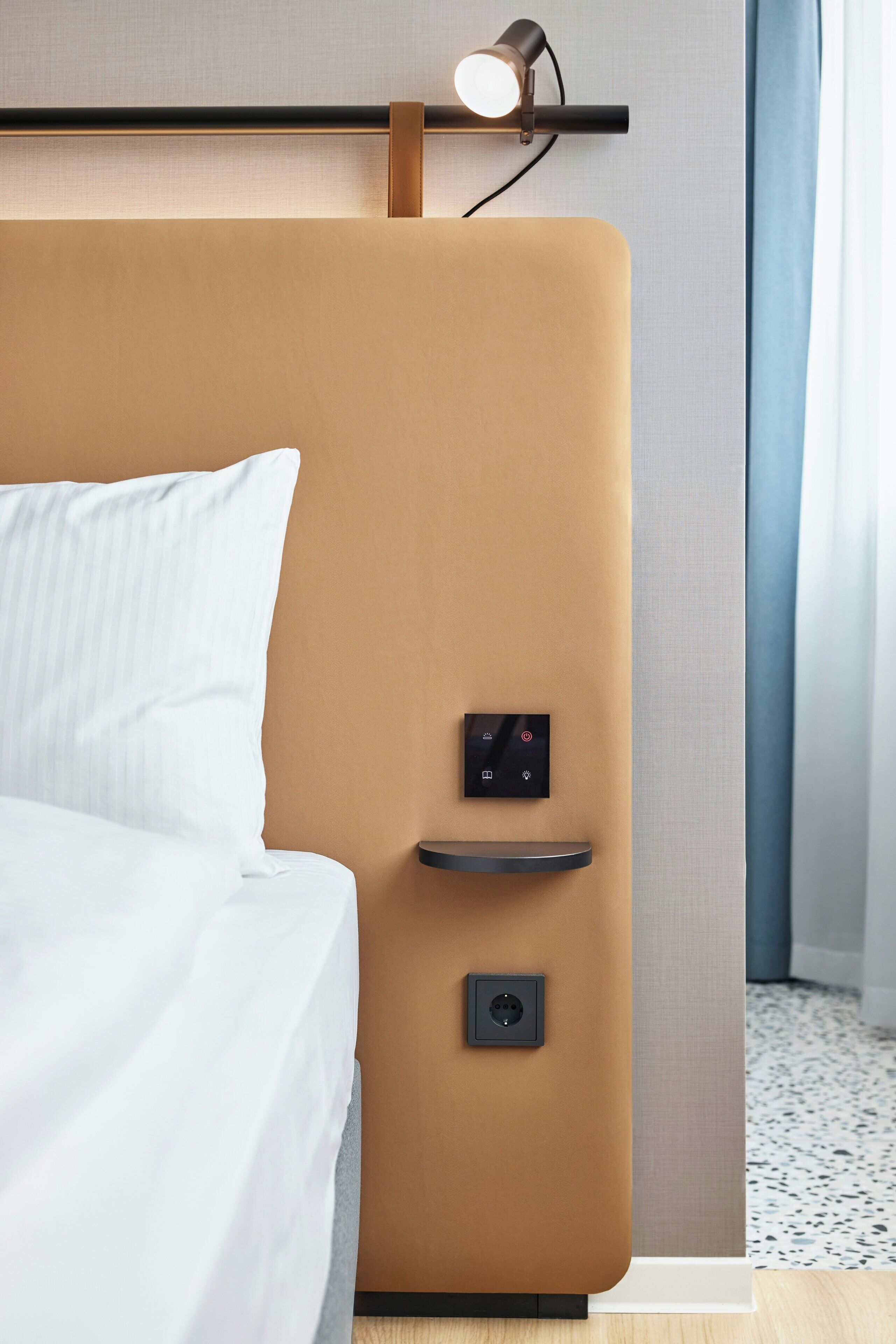 Una habitación de hotel moderna con una cama, un panel de control y una lámpara fijada a la pared.