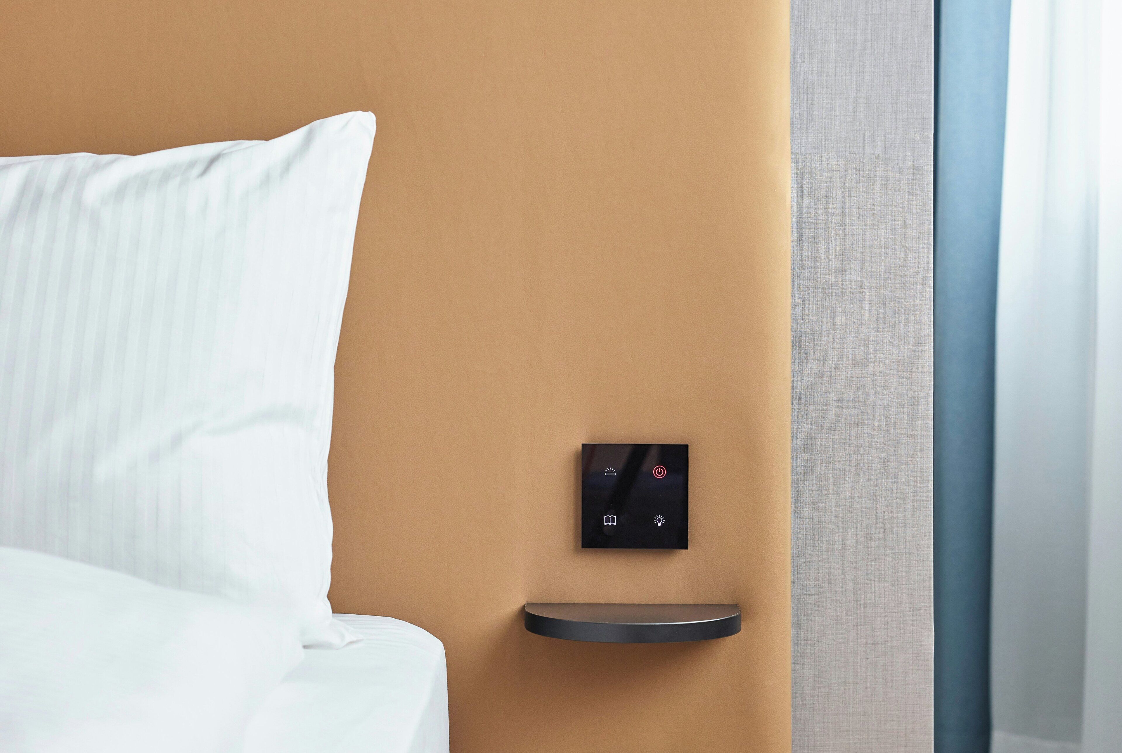 Una habitación de hotel con una cama, un panel de control en la pared y una pequeña estantería.