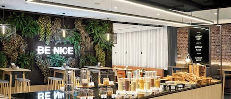 Ein modernes Café-Interieur mit einer zentralen Theke, die Speisen präsentiert, einer Wand mit Pflanzen und dem Schriftzug 'BE NICE' sowie einer Speisekarte rechts.