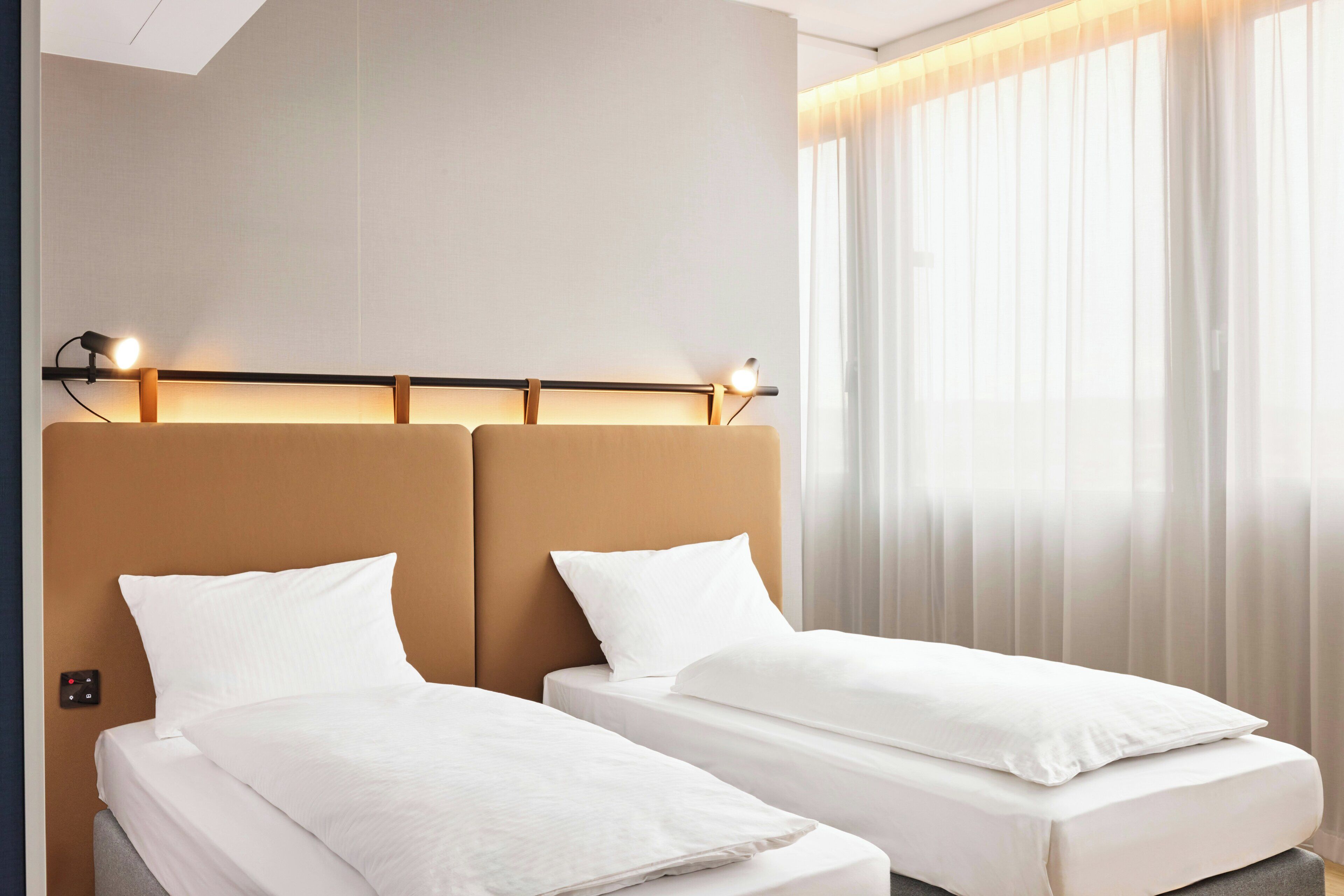 Ein Hotelzimmer mit zwei Einzelbetten, einem Kopfteil mit integrierter Beleuchtung und einem großen Fenster mit durchsichtigen Vorhängen.