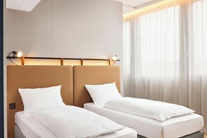 Une chambre d’hôtel avec deux lits simples, une tête de lit dotée d’un éclairage intégré et une grande fenêtre avec des rideaux légers.