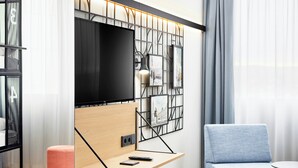 A modern living room with a flat-screen TV mounted on a wall, a wooden shelf with a remote control, a blue sofa, and a yellow ottoman.