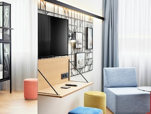 A modern living room with a flat-screen TV mounted on a wall, a wooden shelf with a remote control, a blue sofa, and a yellow ottoman. - Hotel Wien-Schönbrunn (Vienna)