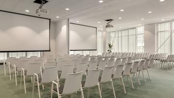 Uma sala de conferências com fileiras de cadeiras brancas, uma tela grande e um projetor fixado no teto.