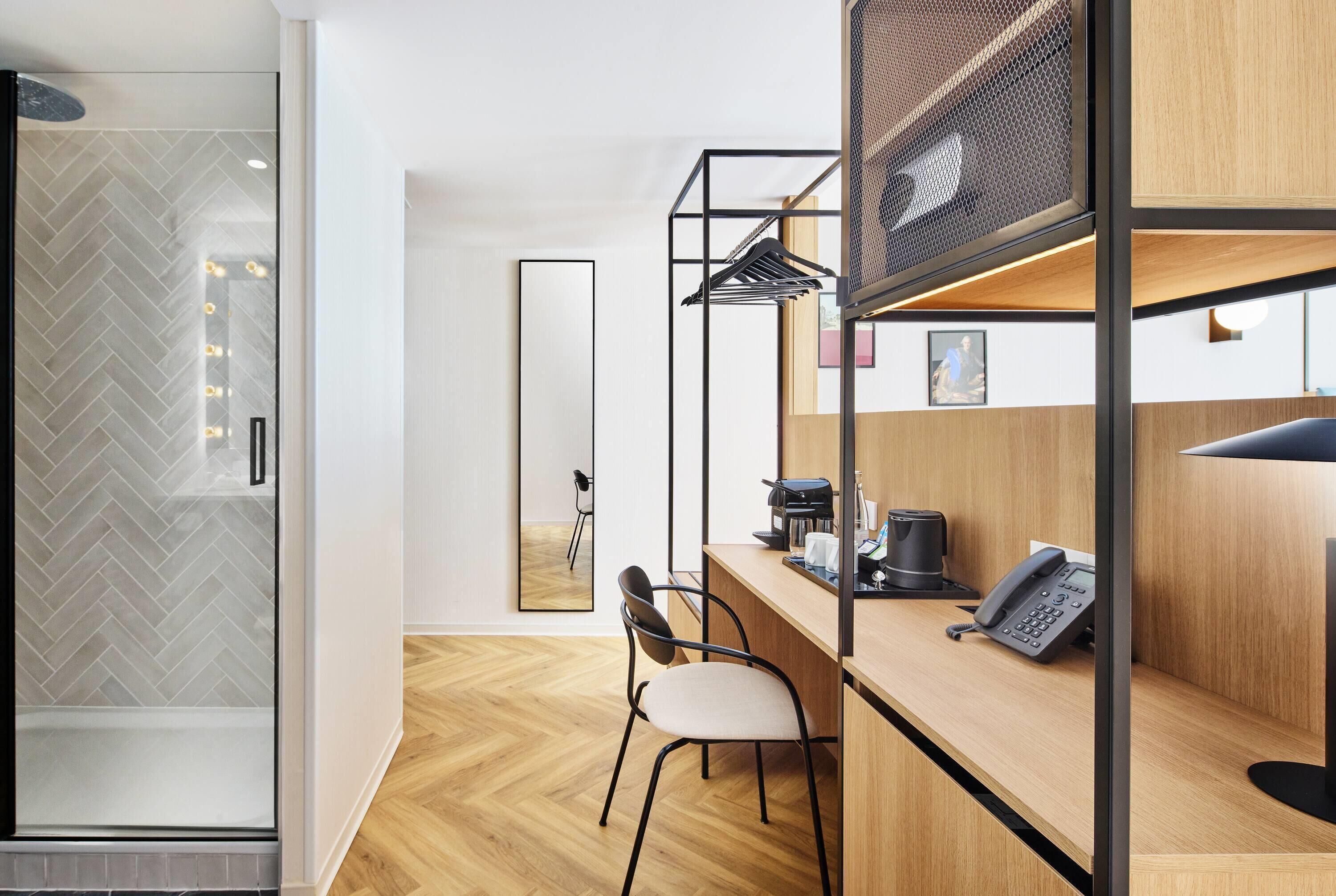 Een moderne hotelkamer met een doucheruimte in visgraatmotief, een houten vloer, een bureau met een koffieapparaat, een telefoon en een spiegel.