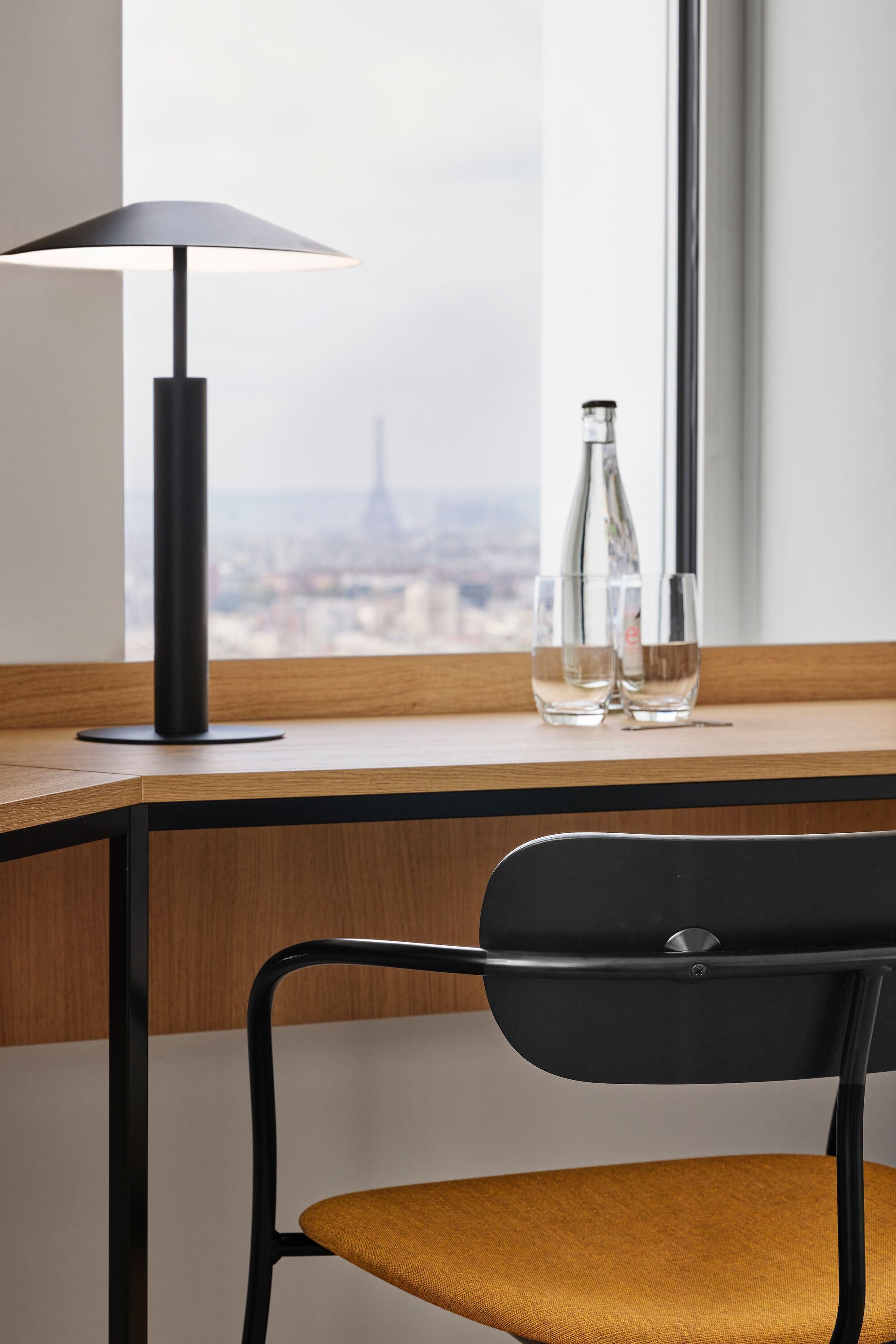 A modern desk with a lamp, two glasses, and a bottle, positioned by a window overlooking a cityscape.