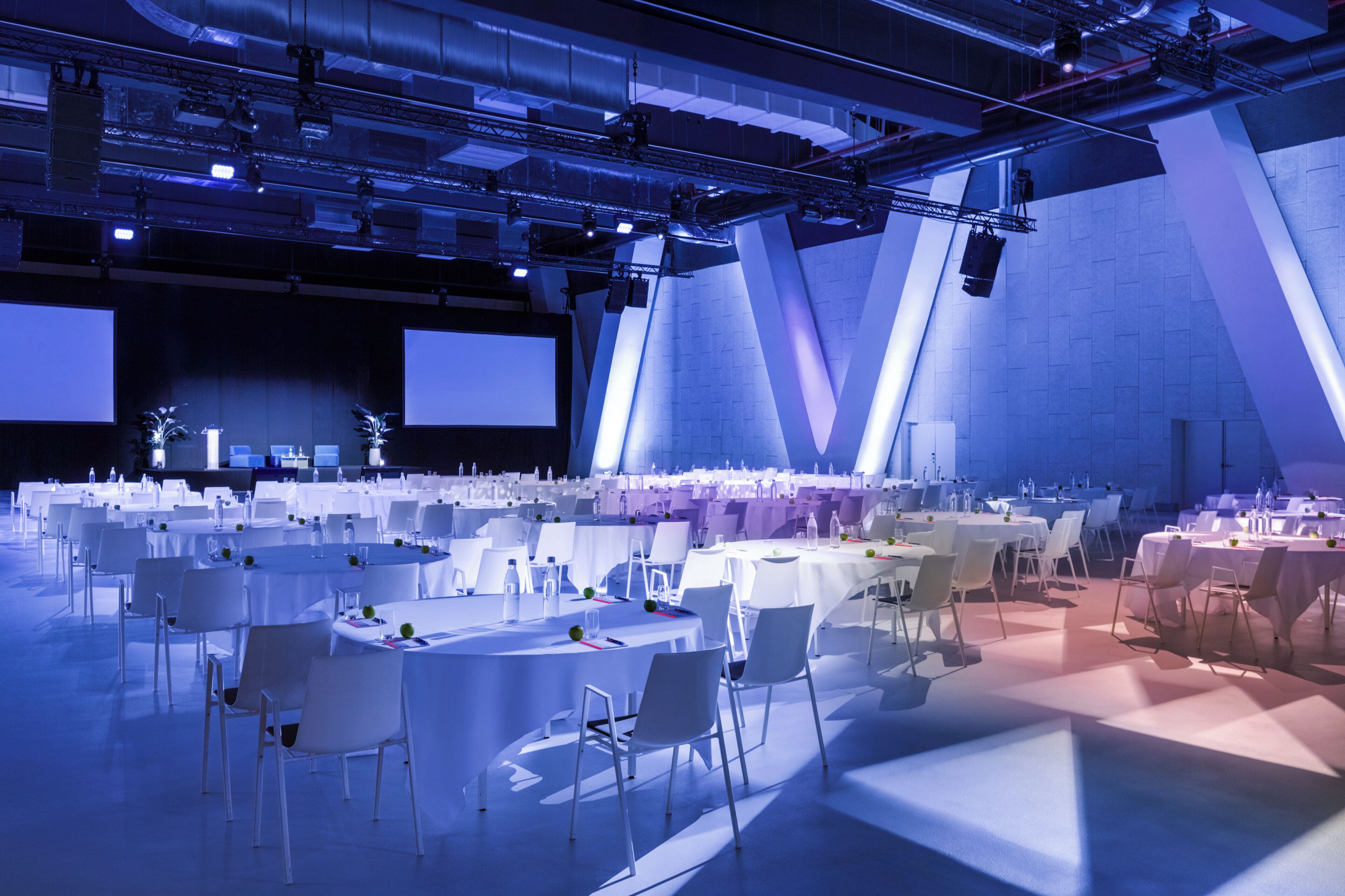 Une grande salle de conférence dotée de plusieurs tables rondes et de chaises, et équipée de projecteurs et d’un système d’éclairage pour les présentations.