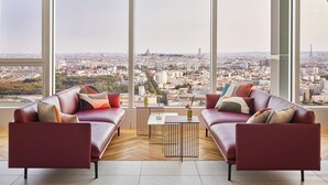 A modern living room with a city view, featuring two leather sofas, a coffee table, and a vase with flowers. - H4 Wyndham Paris Pleyel (Saint-Denis)