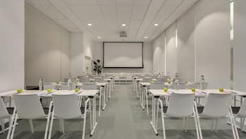 A conference room with white tables, chairs, and a large screen at the front.