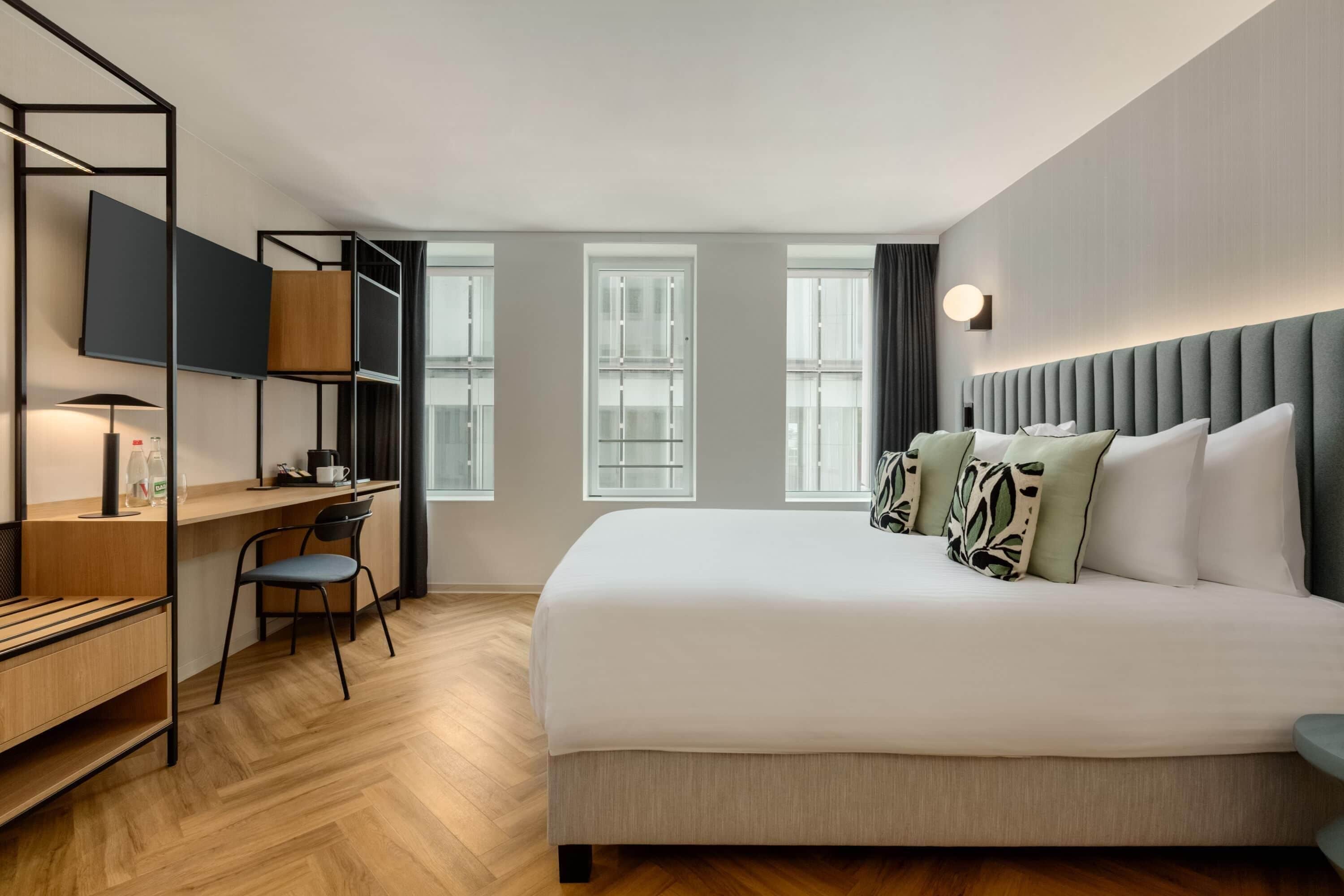 Habitación de hotel con cama, escritorio, silla, televisor y ventanas amplias.