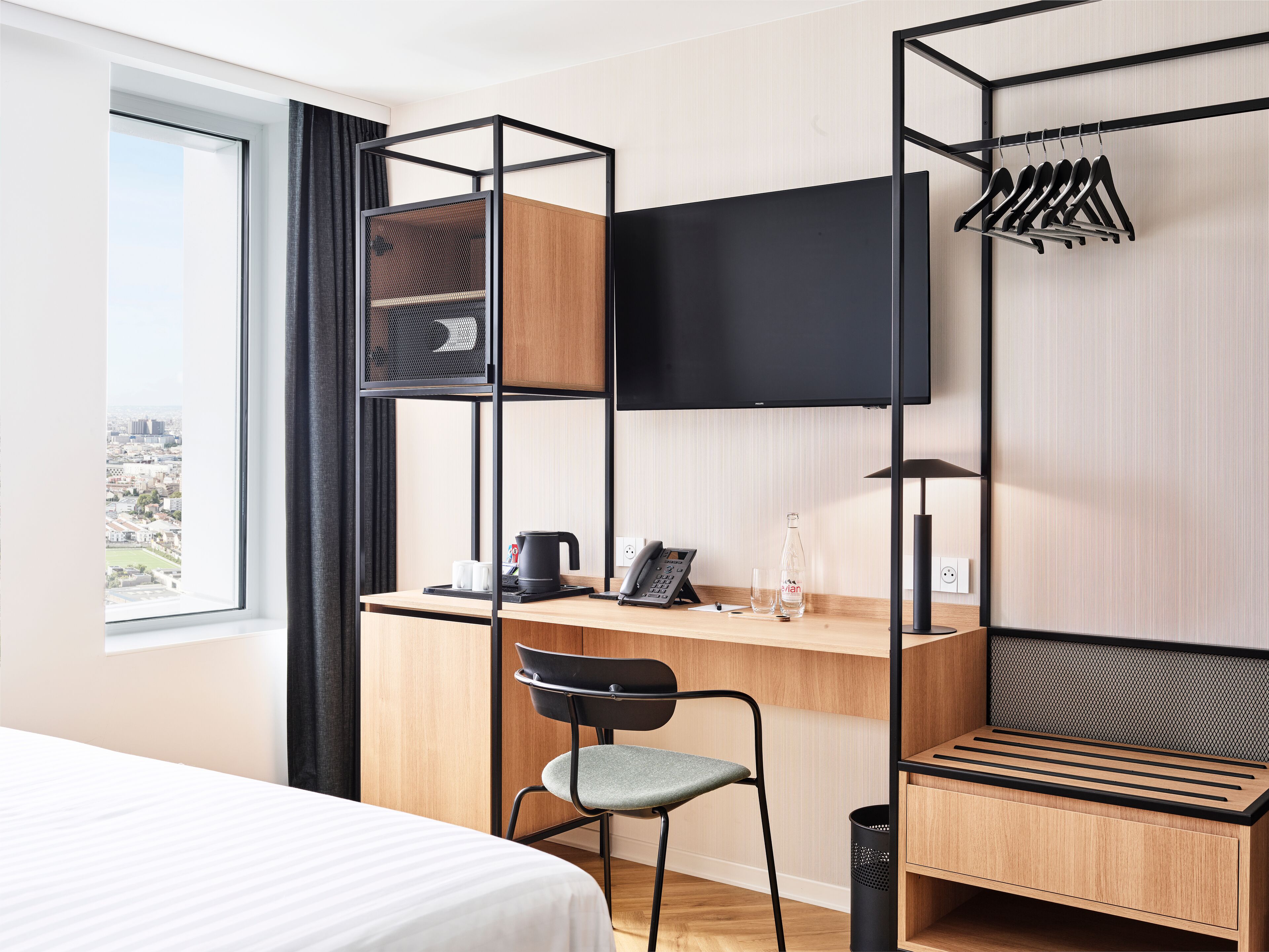 A hotel room with a bed, a desk with a chair, a TV, a shelf with a kettle, a bottle, and a trash bin.