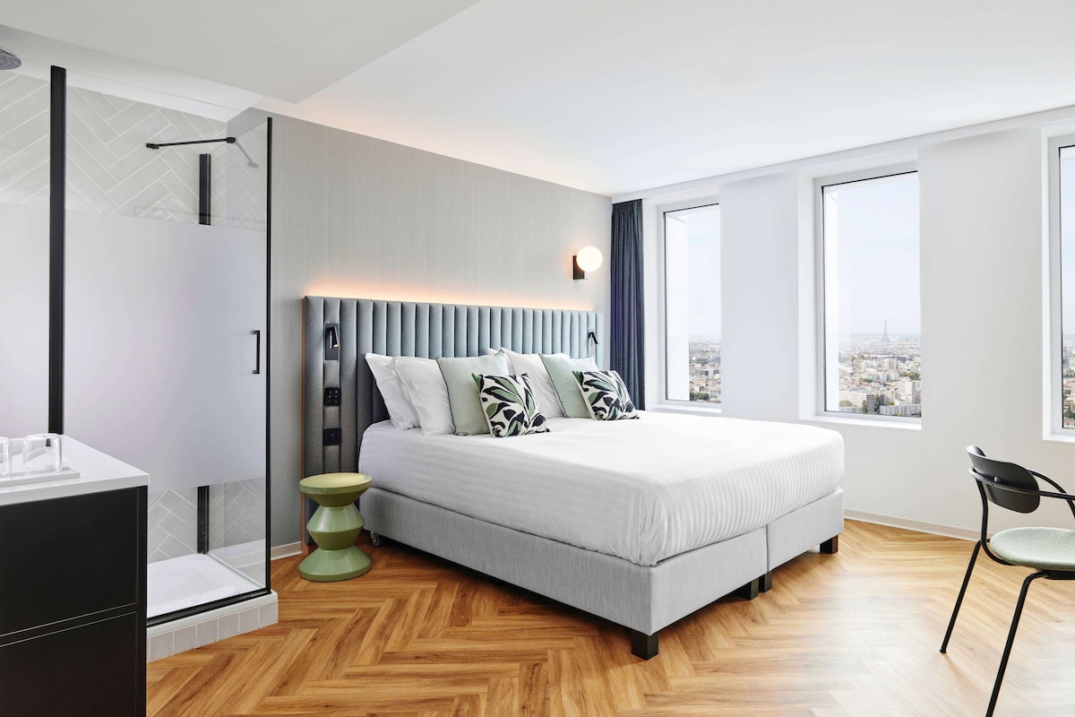 A modern bedroom with a large bed, a glass shower enclosure, and a city view.