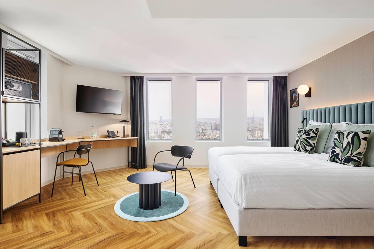 A hotel room with a bed, desk, chairs, a TV, and large windows with a city view.