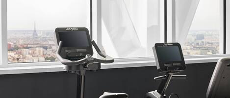Two stationary bikes with digital displays, positioned in front of a window with a cityscape view.