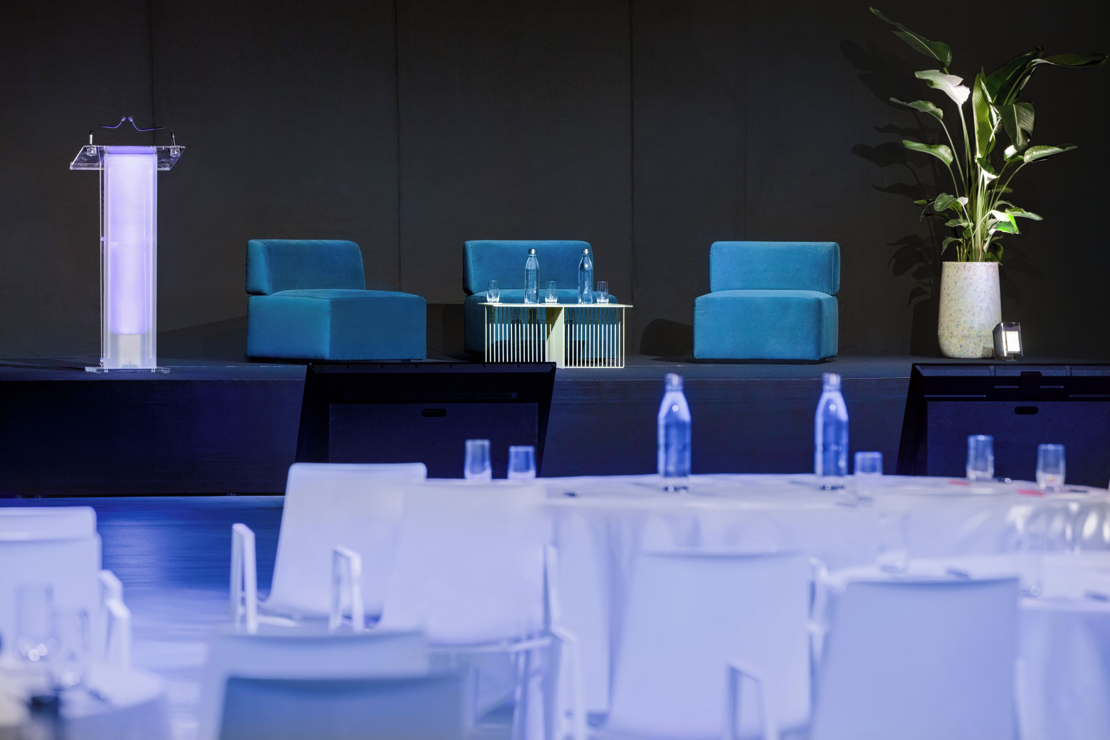 Un salón de conferencias con un atril, dos sillas azules, una mesa con botellas de agua y una planta en maceta.
