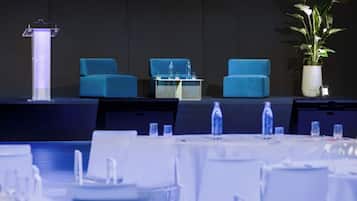 Un salón de conferencias con un atril, dos sillas azules, una mesa con botellas de agua y una planta en maceta.