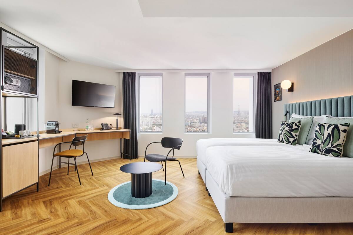 A modern hotel room with a large bed, a desk, two chairs, a television, and a city view.