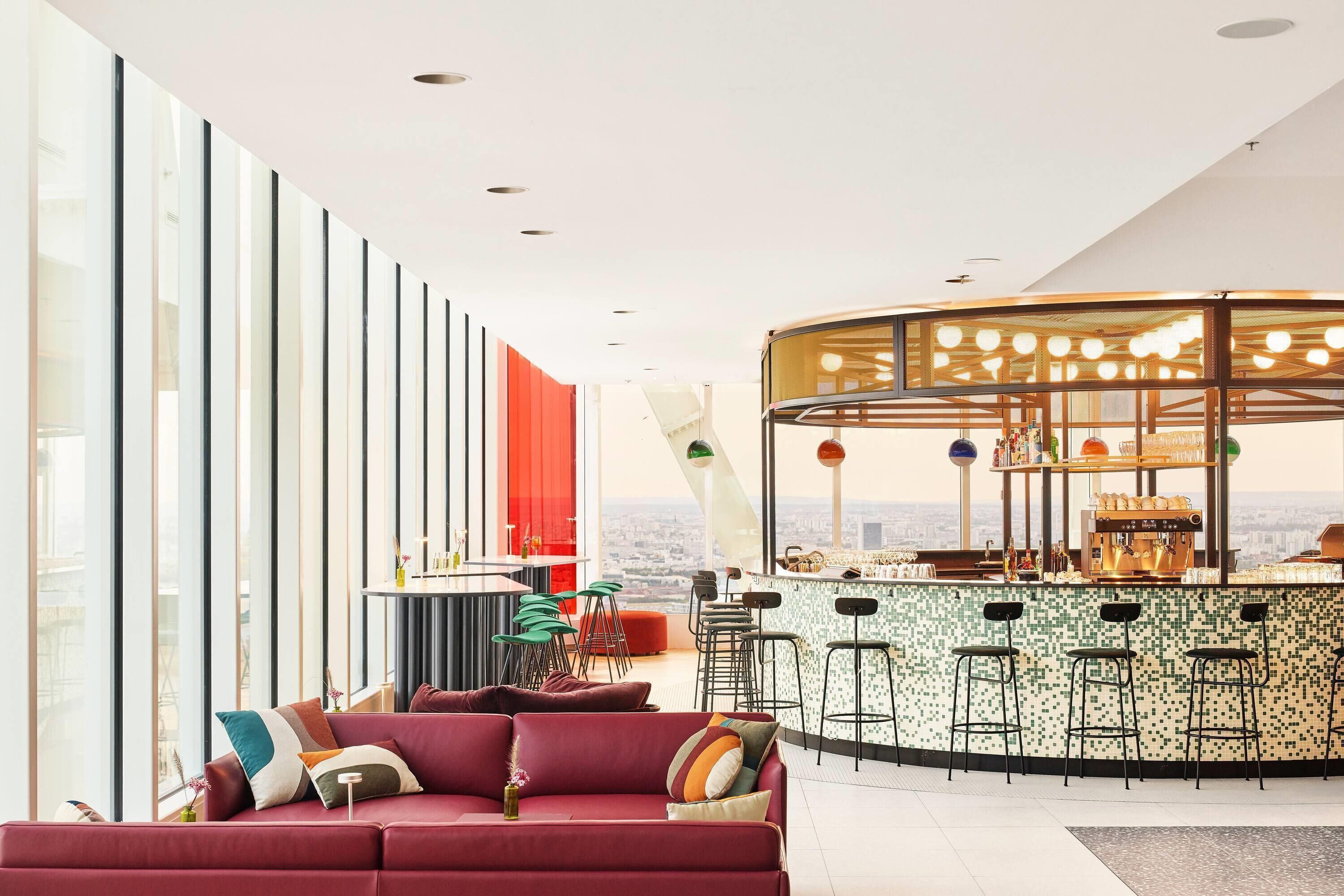 A modern bar area with a city view, a red sofa with cushions, and a bar counter with stools.