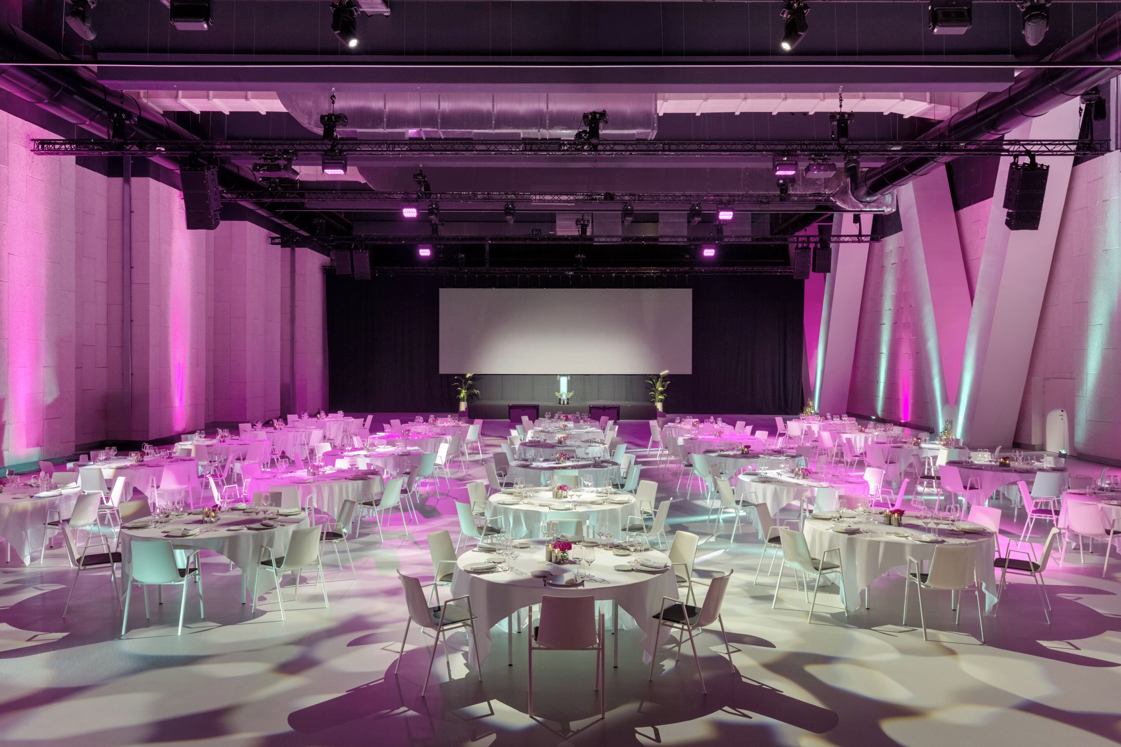 A large conference room with multiple round tables, white chairs, and a stage with a large screen.