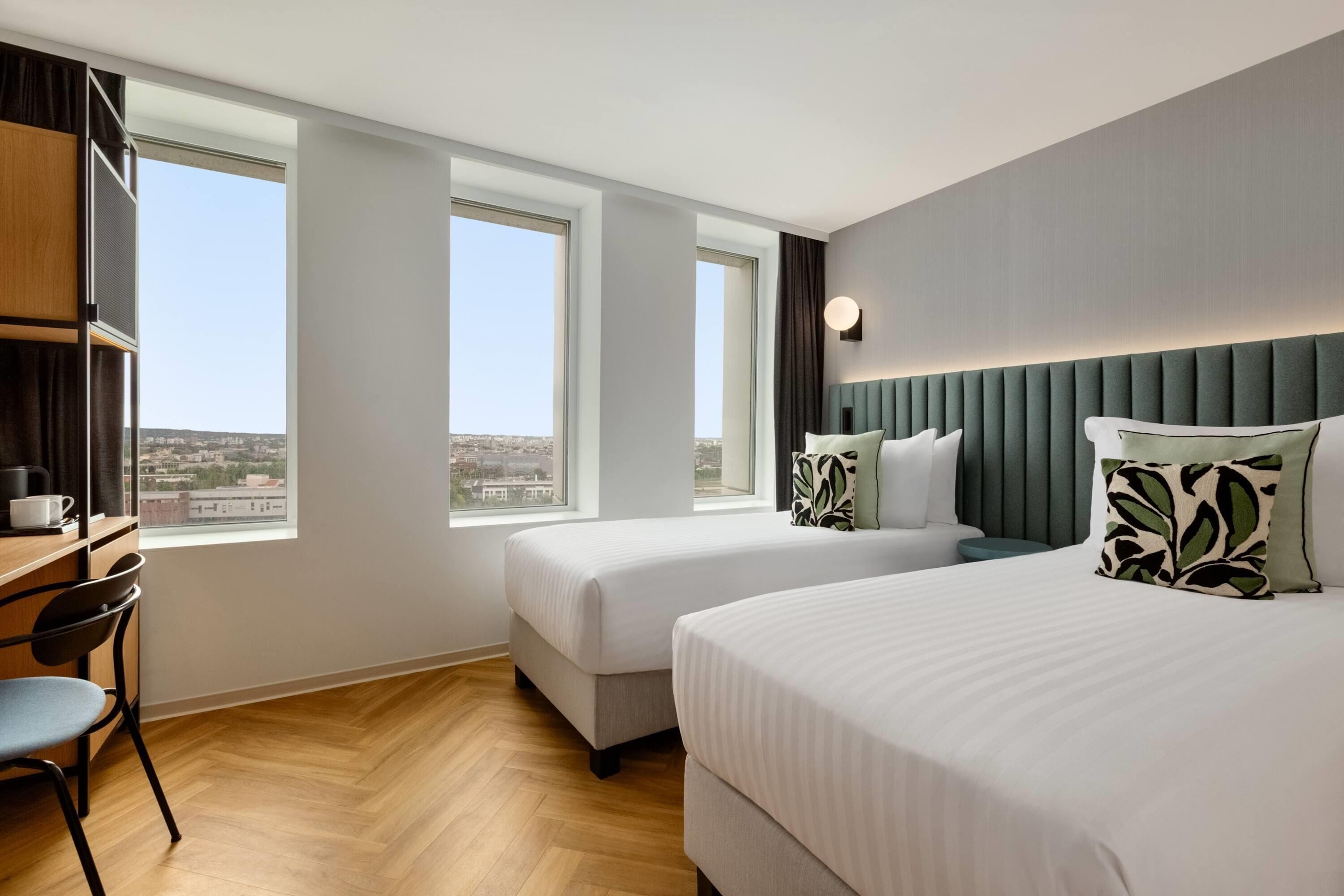 A hotel room with two beds, a desk, and large windows offering a city view.