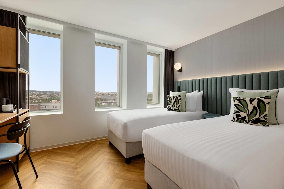A hotel room with two beds, a desk, and large windows offering a city view.