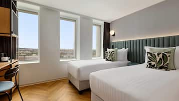 A hotel room with two beds, a desk, and large windows offering a city view.