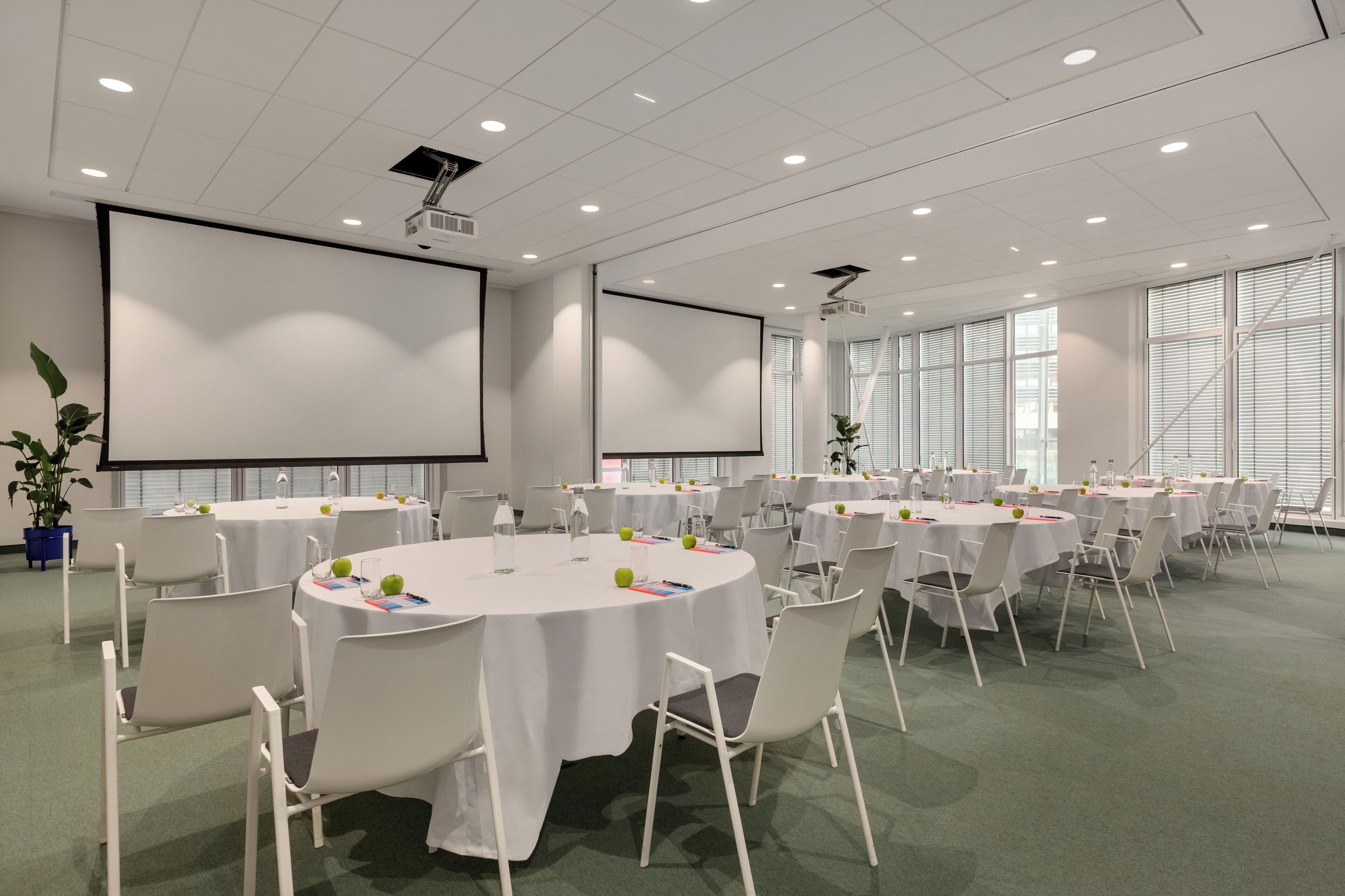 A conference room with multiple tables, chairs, a projector screen, and large windows.