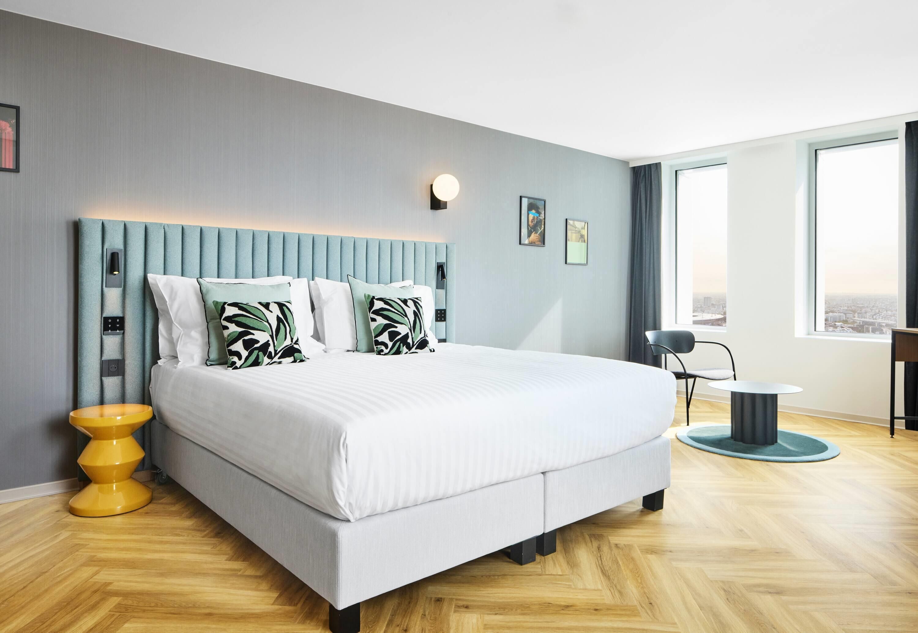 A modern hotel room with a large bed, a bedside table, a chair, a round table, and a view of the city through the window.