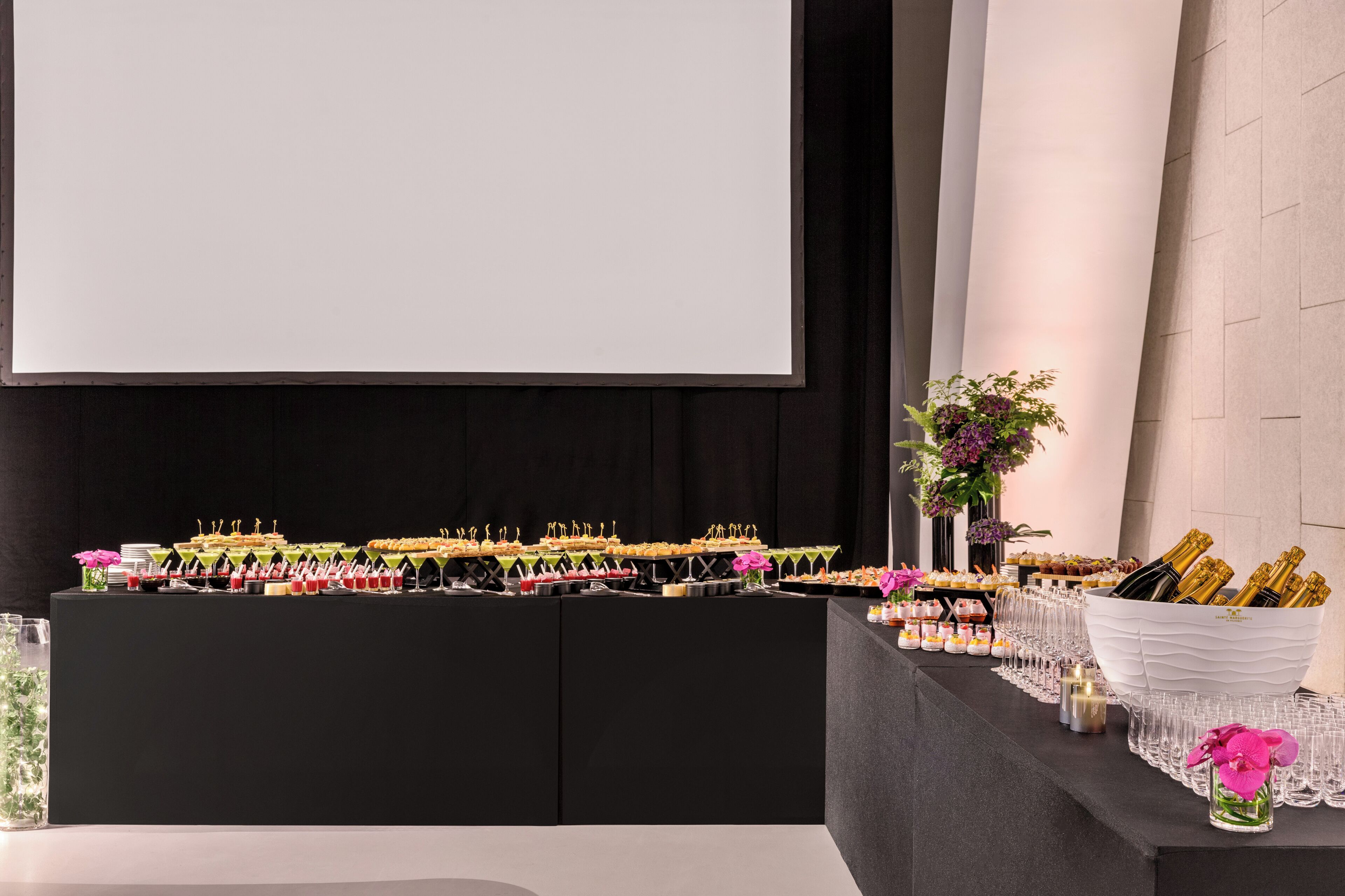 Een buffettafel met diverse gerechten en drankjes, een projectiescherm en een bloemstuk.